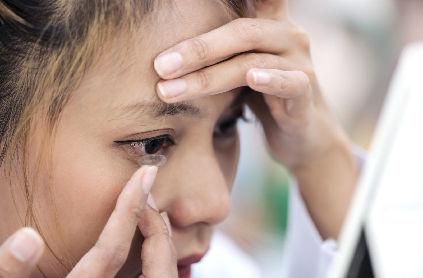 A woman placing a contact lens onto her right eye with her right hand.
