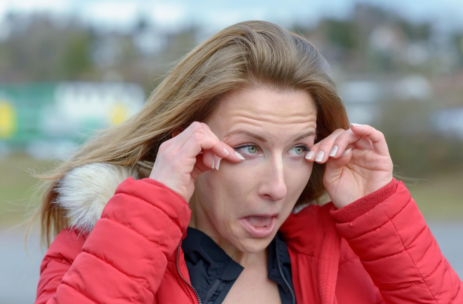 Woman in a red winter coat rubbing her watery eyes outdoors on a chilly day