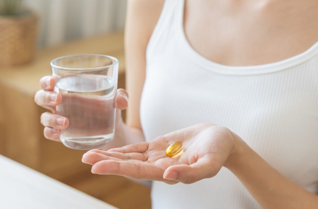 A close-up of a young adult holding a glass of water and a single yellow supplement vitamin to prevent dry eyes.