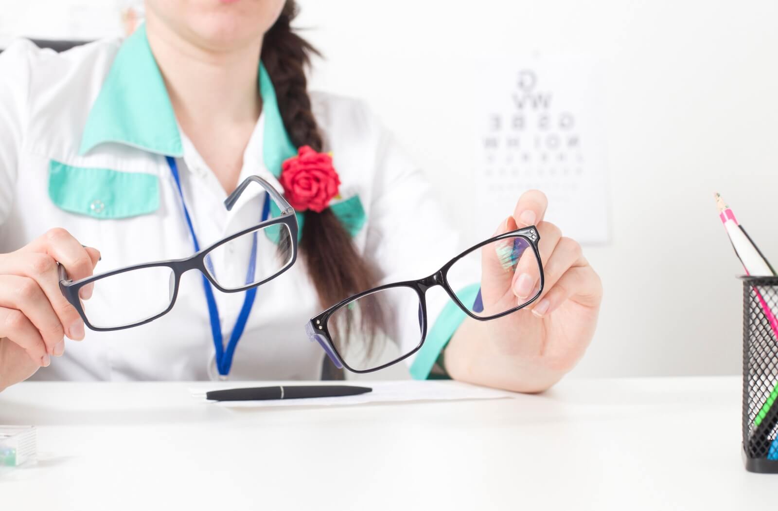 An eye doctor sitting at a white desk and holding different glasses in each hand.
