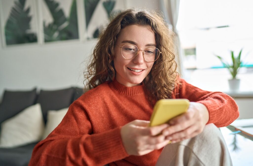 A person wearing glasses and a red sweater smiles while looking at a yellow smartphone.