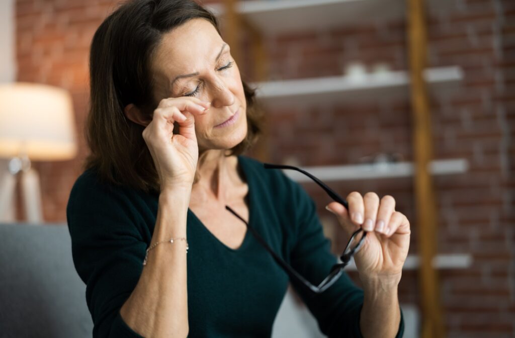 A middle-aged woman removing her eyeglasses to rub her closed eyes in a home setting.
